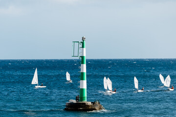 Green and white striped lighthouse stands on rocky shore, guiding small sailboats navigating the calm blue waters, showcasing maritime safety and leisure activities