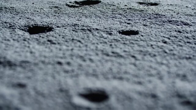 A close-up, grey, rough surface with small craters. The texture appears rocky with dimples