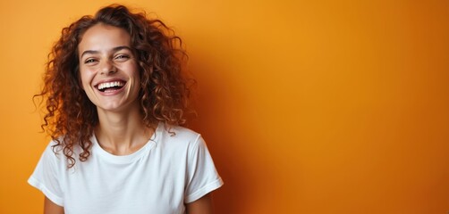 Woman with curly hair laughs joyfully against orange studio backdrop. She wears simple white t-shirt and expresses happiness and confidence. This image conveys positive emotions and self-assurance.