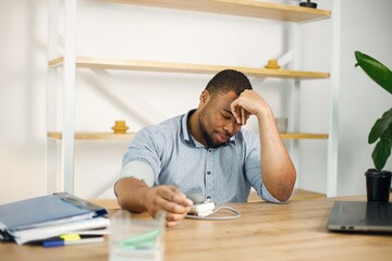 Black male entrepreneur sitting in office near laptop and using tonometr