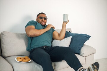 Black guy sitting on a couch in living room drinking a tea and eating a cookies
