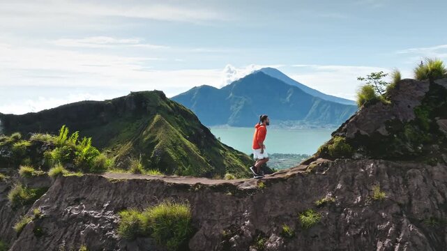 Man hiking a challenging, rocky trail on Mount Batur in Bali, enjoying adventure travel with stunning views of volcanic lake and distant mountains under blue sky. Active sports outdoor. Drone flight