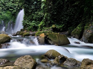 Rushing river water sweeps past mossy stones beneath towering jungle cliffs, blending motion and texture in a vibrant scene of untamed rainforest energy.