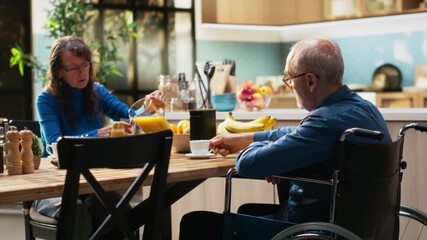 Elderly man wheelchair user drinking coffee at breakfast with his wife, smiling and chatting together in a relaxed morning routine. Peaceful retired lifestyle at home, kitchen table. Camera B. - Powered by Adobe