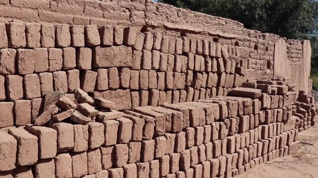 Stacked handmade adobe bricks on arid ground against a traditional mud wall. Dry and sunny environment in Fiambala Catamarca.