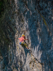 Obraz premium LOW ANGLE VIEW: Male lead climber works his way up a steep limestone route, gripping rock while sunlight casts shadows across the wall. His rope trails below as he climbs in natural climbing area.