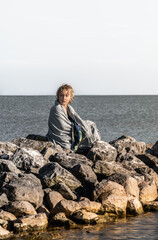 A young boy sits on large boulders by the sea, wrapped in a towel, looking over his shoulder with a neutral expression. Water surrounds the boulders under a clear sky.
