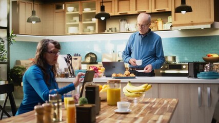 Elderly male adult browsing online on laptop in rustic kitchen space, enjoying cozy moment during morning routine with coffee. Peaceful retired life and wellbeing for happy lifestyle. Camera A.