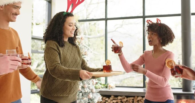 Mid adult woman in green sweater offering mulled drinks and friends clinking glasses in living room