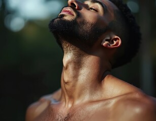 Bearded man tilts head up towards sunlight. Muscular male relaxes in nature outdoors. Adult person poses confidently, enjoying summer day in natural green environment.