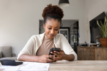 Positive young African American woman using online financial application, Internet bank service on mobile phone, typing on smartphone at laptop with heap of bills and calculator