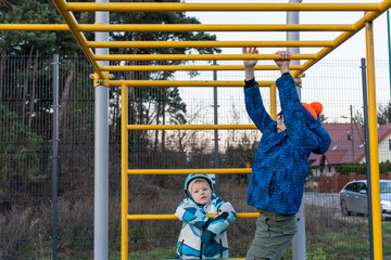 Two brothers at outdoor calisthenics park, older boy hanging on monkey bars while younger child in helmet watches on autumn day. Concept of siblings playing together, street workout and active family.