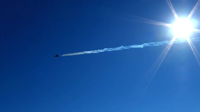 A powerful aircraft streaks across an endless vibrant blue sky, leaving a prominent contrail under the bright sun's glare