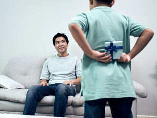 Son hiding gift behind their back while approaching father at home, portraying heartwarming surprise moment, family bonding and emotional connection