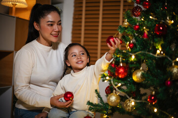 mother and toddler girl decorating Christmas tree in home at night
