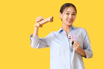 Young Asian woman with driving license and toy car on yellow background