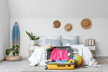 Interior of light bedroom with cozy bed, surfing board and men's swimwear in open suitcase