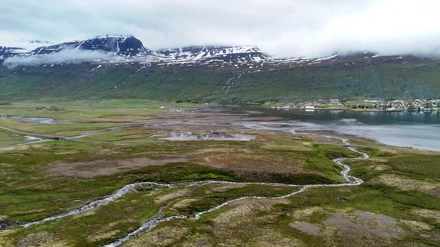 Wide aerial view of the mountains near F&aacute;skr&uacute;&eth;sfj&ouml;r&eth;ur, with snow-lined peaks, scattered waterfalls, winding wetlands, and an open valley crossed by a curving road under cloudy skies