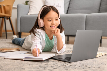 Little Asian girl in headphones with notebook and laptop studying online at home
