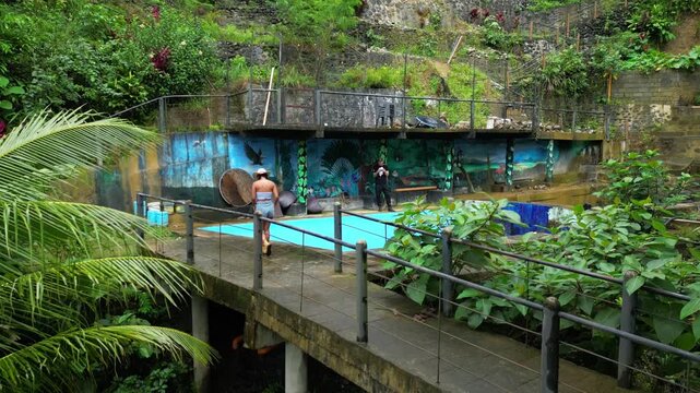 Forward aerial surpassing woman walking elevated concrete path with metal railings, revealing lush tropical foliage, palm trees, and poolside mural in Tanay Rizal, Philippines.