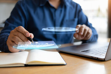 A person using a pen to write in a notebook with a laptop on the desk and a holographic display showing a bar chart and upward arrow above the notebook