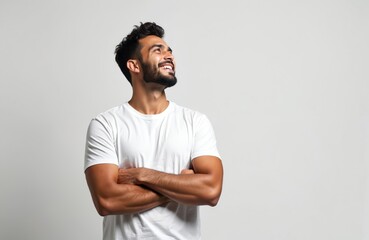 Young man stands with arms crossed looking up and smiling. He appears happy confident and hopeful. Man has dark hair and beard. Clean studio background provides copy space.