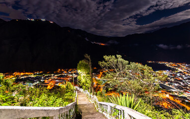 Illuminated stone stairs of Mirador de la Virgen Trail overlook the glowing city lights of Banos de Agua Santa, Ecuador. Night landscape features Andean valley and dark mountains
