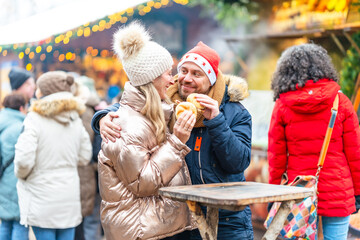 Smiling couple eating warm sausages and rolls at a Christmas market surrounded by glowing festive lights