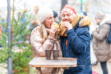 Happy couple enjoying tasty sausages and rolls at a Christmas market surrounded by warm seasonal lights