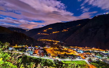 Illuminated winding road of Via a Runtun rises above Banos de Agua Santa at night in Ecuador. Scenic landscape features glowing city lights and dark Andean mountains