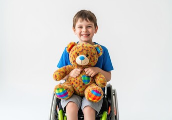 Happy young boy with disability sitting in a wheelchair and hugging his bright teddy bear toy.