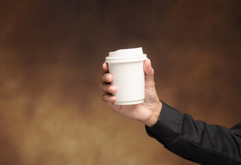 Hand holding white paper coffee cup on brown background.