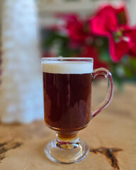 A festive glass mug filled with a dark hot beverage and topped with foam, placed on a wooden table with soft holiday décor in the blurred background. Warm, cozy seasonal atmosphere.