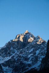 Extreme close-up of a rugged, rocky mountain peak dusted with snow, dramatically lit by low winter sun against a clear blue sky