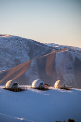 Three dome-shaped glamping accommodations situated on wooden platforms in a snow-covered valley beneath arid, snowy mountain slopes