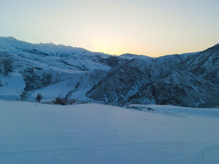 Fototapeta premium Wide aerial view of steep, snow-dusted mountains meeting a large, flat snow-covered field with a single tree and distant structures visible under a sunrise sky