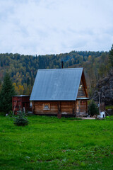A simple wooden cabin with a gray metal roof stands in a vibrant green meadow against a backdrop of dense, mixed forest covering a steep hillside under a cloudy sky