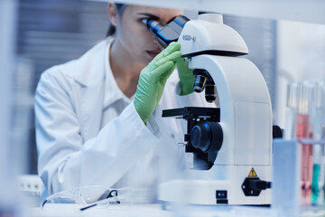 Caucasian young adult woman examining sample using laboratory microscope, wearing protective gloves and lab coat, focusing on scientific research in clinical laboratory setting