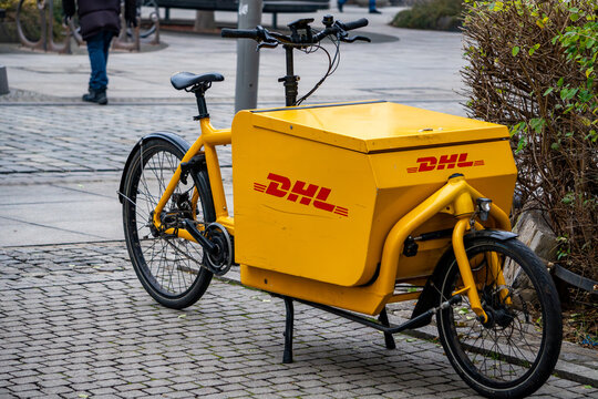 Yellow DHL cargo bike with large transport box positioned on urban cobblestones surrounded by greenery and pedestrian walkway elements