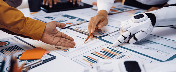Professionals engage in a collaborative effort, using both human and robotic hands to analyze detailed business charts in a modern office environment, emphasizing innovation.Noogenesis