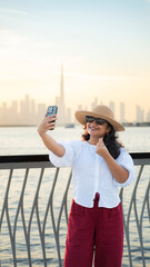 Fototapeta premium Solo traveler taking selfie with Dubai skyline at sunset standing by waterfront deck enjoying iconic architecture and city views