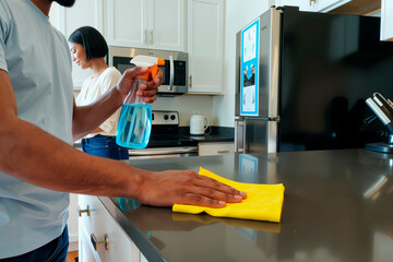 Young adult man cleaning kitchen counter with spray bottle and yellow cloth while young adult woman standing in background near stove preparing food