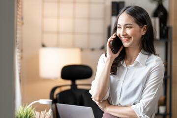 Modern Asian businesswoman smiling and having a professional phone call while standing in a bright, contemporary office environment.