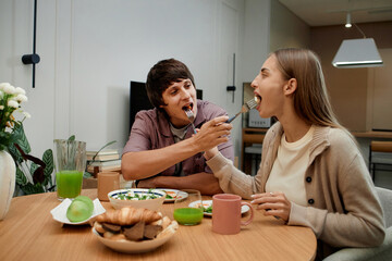 Young Caucasian man feeding young Caucasian woman with fork while sitting together at dining table, both smiling and enjoying breakfast with croissants and green juice