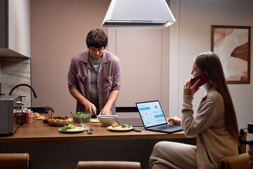 Caucasian young adult man preparing food at kitchen counter while Caucasian young adult woman sitting nearby multitasking with laptop and smartphone, both engaged in daily routine
