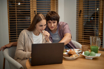 Caucasian young woman and Caucasian young man sitting at table using laptop together, woman eating snack while man pointing at screen, breakfast food and drinks on table