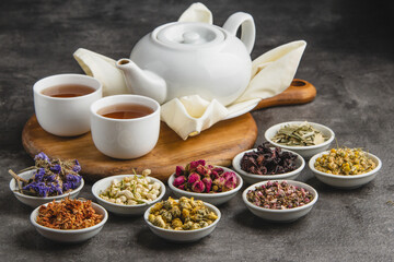 A white teapot and two cups of tea surrounded by small bowls of various dried herbs and flowers for herbal infusions on a dark background.