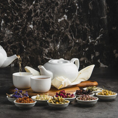 A white teapot pouring hot herbal tea into a cup, surrounded by small bowls of dried flowers and herbs for infusion.