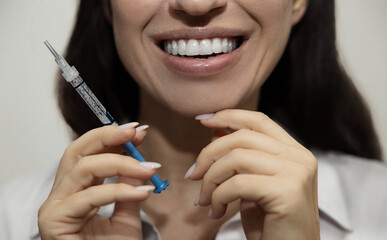Woman holds whitening trays and hydrogen peroxide syringes, smiling while showing a dental model. White backdrop, close frames with trays, gel and tooth care tools for a bright healthy smile.