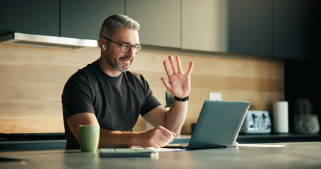 Laptop, remote work and video call with man in kitchen of home for planning, report or research. Computer, earphones and wave with mature freelancer in apartment for virtual meeting or webinar